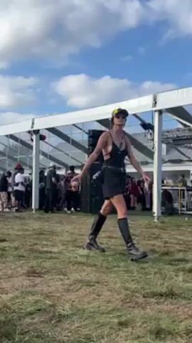 The video shows a music festival with a DJ booth set up under a large white tent on a grassy field. People are gathered in front of the booth, some dancing and others milling about. A woman in a black bra top, black shorts, and high boots is standing in the foreground, moving slightly. The sky is partly cloudy. A stack of large black speakers sits next to the tent.