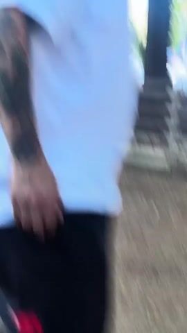 A medium shot shows a man with tattoos on his left arm, wearing a white shirt. The camera then pans to the left, showing a wooden fence in the foreground. In the background, there are trees, people, and several tents. The camera then pans back to a close-up of the man's face, showing his dark hair, a small goatee, and a lip piercing.