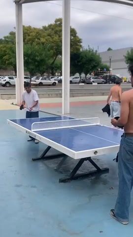 Two young men are playing ping pong outdoors. The man on the left is wearing a white t-shirt and blue jeans and is holding a red ping pong paddle. He is facing the table and appears to be mid-swing. The man on the right is shirtless, wearing blue jeans, and is facing away from the camera, holding a ping pong paddle. He is standing behind the table. The ping pong table is blue with a white border and a white net. The court is blue asphalt with some puddles of water on the surface, suggesting it may have recently rained. In the background, there are trees, parked cars, and buildings under a cloudy sky. The scene is under a white awning supported by white pillars.