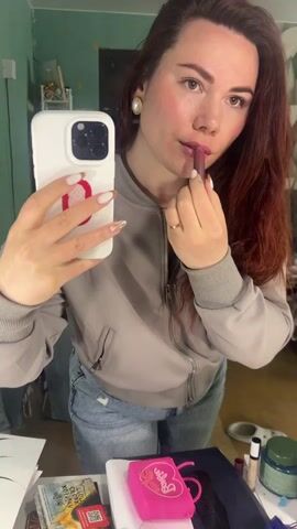 A woman is applying lipstick while holding a smartphone in her left hand, capturing a selfie video. She is wearing a light grey bomber jacket and blue jeans. Her hair is long and reddish-brown. She has white and gold nail polish with designs. She is wearing pearl earrings. Behind her is a cluttered space with various items on a table, including a book, a pink Barbie purse, and other cosmetic products. The walls are a pale green color.