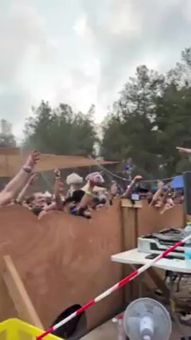The video shows a crowd of people dancing at an outdoor event. Many people are shirtless or wearing swimwear. They appear to be enjoying themselves and dancing to music. A wooden structure is visible in the foreground, possibly a makeshift DJ booth or stage. There's a large tent or shade structure overhead. The setting appears to be a wooded area with trees in the background and a cloudy sky above. Some are filming with their phones.