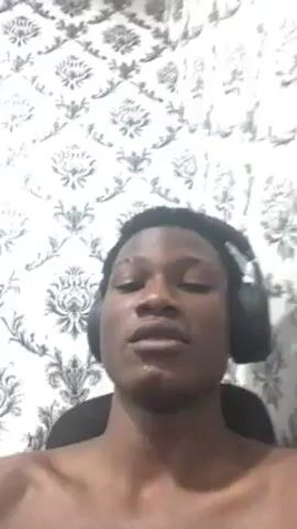 A dark-skinned person wearing headphones is singing. The person is topless and seated in front of a white and gray floral patterned wall. The camera is pointed up toward the person's face. They move their head and mouth as they sing.