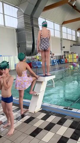 An indoor swimming pool facility features several children and adults. In the foreground, a girl wearing a black and white checkered swimsuit and a green swim cap jumps into the pool from a white diving board, creating a splash. Another girl, wearing a purple and white patterned swimsuit and a green swim cap, steps onto the diving board, preparing for her turn. A boy in blue swim trunks and a green swim cap stands nearby, watching. His swim cap has "PETITS PLOUF" and "ETIERS" written on it.

The pool area has a black and white checkered tiled floor leading up to the pool edge. In the background, an adult male stands near the pool, and another adult is seated on a tall lifeguard chair. Various swimming aids like floats and kickboards are visible in racks beyond the pool. The facility has a high ceiling with large industrial-style ventilation ducts and large windows providing natural light. The water in the pool is turquoise.