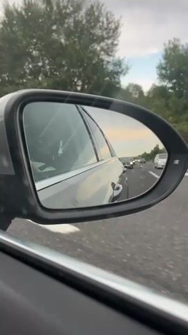 The video is a first-person perspective shot from inside a car, focusing on the side-view mirror. The mirror reflects a highway with a line of cars in the distance. Trees and the sky, with hints of a sunset, are visible through the car window in the background. The road appears to be multi-lane with a white line marking the edge. The car is moving, indicated by the blurred trees and the reflection of the road.