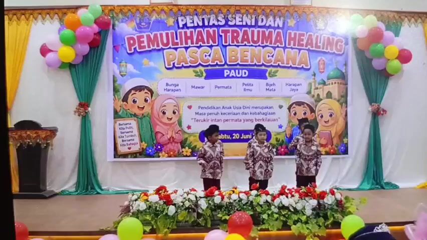 The video shows a stage performance at an event for young children. A large banner in the background reads "PENTAS SENI DAN PEMULIHAN TRAUMA HEALING PASCA BENCANA PAUD," which translates to "Art and Trauma Healing Performance Post-Disaster PAUD." PAUD likely refers to early childhood education. The banner features cartoon drawings of children in Islamic attire, mosques, and balloons. Below the title, there are labels such as "Bunga Harapan" (Flower of Hope), "Mawar" (Rose), "Permata" (Jewel), "Pelita Ilmu" (Light of Knowledge), "Bijih Mata" (Seed of the Eye), and "Harapan Jaya" (Great Hope). A quote in Indonesian is also visible: "Pendidikan Anak Usia Dini merupakan masa penuh keceriaan dan kebahagiaan. Terukir indah permata yang berkilauan" (Early Childhood Education is a time full of joy and happiness. A beautiful, sparkling jewel is engraved). The date "20 Juni 2020" is displayed.

On the stage, three young boys in matching traditional patterned outfits are standing in front of a floral arrangement. They appear to be performing or about to perform. The stage is decorated with green drapes and balloons in various colors, including red, yellow, green, and pink. There are also additional floral arrangements and some balloons on the floor in front of the stage. The lighting appears to be artificial, illuminating the stage. The environment suggests an indoor event, possibly a school or community hall.