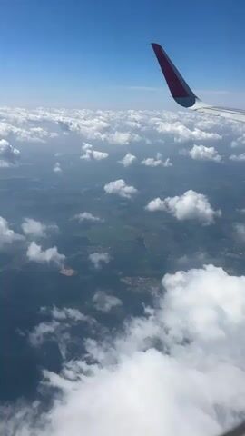 The video provides an aerial view from an airplane window, looking out at a landscape dotted with clouds. The primary subjects are the clouds, the airplane's wing, and the land below. The scene is set during the daytime, with a bright blue sky and sunlight illuminating the clouds, suggesting fair weather.

The land below appears to be a rural or semi-rural area, with visible fields, patches of trees, and scattered settlements or buildings. There are distinct patterns of agricultural land and perhaps some roads or waterways winding through the terrain. The clouds are white and fluffy, some are smaller and scattered, while others form larger, denser formations, casting shadows on the ground.

The airplane wing, with a red tip, is a prominent object in the frame, indicating the perspective of someone traveling by air. The video captures the activity of flying, showcasing the view from cruising altitude.

While there is no visible text in the video, the location context provided, Nikol'skoye, Russia, allows for potential inferences about the geography being viewed, though specific landmarks are not identifiable from the footage alone. The overall mood conveyed is one of peaceful travel and the beauty of the aerial landscape.