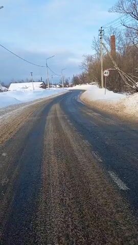 The camera pans across a winter landscape. A road runs through the scene, with patches of ice and snow. On either side, snow covers the ground. In the distance, there are houses and trees. A tall brick smokestack stands out against the blue sky with light clouds. There is also a 40 speed limit sign near the road. Utility poles line the road. The scene appears to be a rural or suburban area during winter.