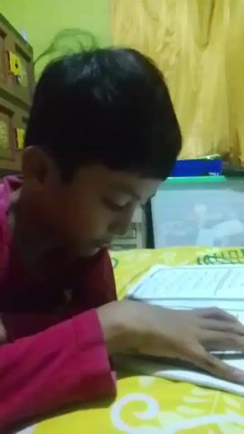 A young boy wearing a pink shirt is seen looking down at a book. The boy's left hand is resting on the book, and his right hand is on the right side of the book. The boy's hair is dark and short. The book appears to have writing on it, with lines and text visible. The background shows a yellow curtain and part of a wooden cabinet. The lighting is dim, with a yellowish hue. The overall scene suggests the boy is engaged in reading or studying.