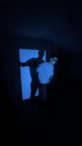 The video shows a man in a blue-lit room dancing in front of a white screen. The lighting creates a stark shadow of him on the screen behind him. He is wearing a white t-shirt, dark pants, and sunglasses. He performs a variety of dance moves. The room is dark and appears to be a small, possibly attic, space. The music is loud and upbeat.