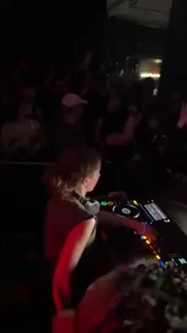 A DJ is playing music at a nightclub or concert venue. The DJ, a young person with their hair in a ponytail, is operating a complex DJ console with multiple knobs, faders, and displays. A crowd of people is visible in the background, illuminated by colored lights. Some of the crowd members are dancing and raising their hands in the air. One person wears an Estonia jersey. The atmosphere is energetic and lively.