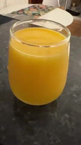 A glass of orange liquid sits on a dark, speckled countertop. The liquid appears to be juice. Behind the glass, a white table with a floral pattern is partially visible, along with white chairs. The overall environment suggests a kitchen or dining area.