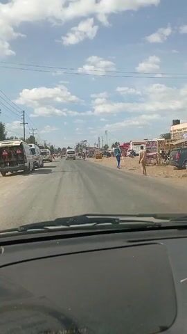 The view is from inside a car driving on a road. On the left, there are a couple of vans and telephone poles. On the right, there are some buildings and a few people walking around. The sky is blue with some scattered clouds. There are a few other cars driving on the road in the distance.