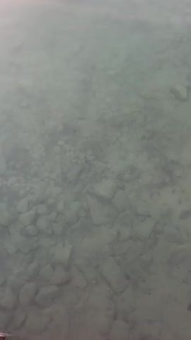 The video begins with a close-up of the shallow bottom of a body of water, revealing rocks and pebbles. As the camera pans up and away, the surface of the water becomes the focus, reflecting a soft, pinkish-orange sunset sky. The horizon is visible, showing a distant, hazy shoreline with rolling hills. The water is calm, with gentle ripples distorting the reflection of the clouds and sky. The color palette transitions from the muted tones of the water's edge to the vibrant hues of the sunset. The video is entirely focused on the natural landscape, with no people or man-made objects visible.