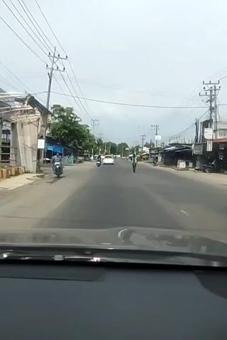 The video is filmed from the perspective of someone in a vehicle, likely a car, looking out through the windshield. The vehicle is driving down a two-lane road in what appears to be a rural or suburban area. On either side of the road, there are businesses and stalls, with some trees and power lines visible. The sky is overcast.

In the distance, a person in a yellow safety vest and dark pants is standing in the middle of the road. They appear to be directing traffic or signaling. Several other vehicles, including motorcycles and cars, are also on the road. The person in the yellow vest is stationary at the beginning of the clip and remains in that position for the duration of the video. No interactions between the person and the vehicles are explicitly shown.