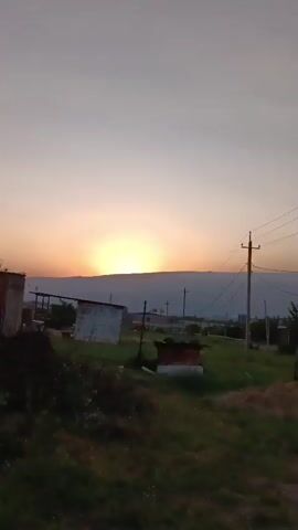 A video captures a sunset over a mountain range. The sun is partially obscured by the mountain, casting a warm orange glow across the sky. In the foreground, there is a building with a white facade and a dark roof. A power pole with wires is visible on the right side of the frame. The overall scene is peaceful and serene, indicative of dusk.