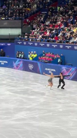 The video shows a pair of figure skaters performing on an ice rink. The male skater is wearing a black outfit, and the female skater is wearing a sparkly dress. They are executing a combination of spins and steps, moving around the ice in sync. The rink is surrounded by a crowd of spectators sitting in the stands. The arena is decorated with the Olympic rings and banners that say "Milano Cortina 2026." The camera follows the skaters' movements, keeping them in frame.