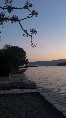The video captures a tranquil sunset or dusk scene over a body of water, likely a bay or large lake, bordered by hills.

At the beginning, a branch with small, dark berries or fruits hangs into the top left of the frame, silhouetted against the twilight sky. Below it, a stone-paved walkway or pier extends along the left edge of the water. A person is briefly visible standing or walking on this walkway on the far left. The water shows slight ripples and reflects the sky's colors.

As the camera slowly pans to the right, the branch moves out of view. The frame opens up to a wide expanse of calm water. In the distance, dark silhouettes of hills or mountains frame the opposite shore. The sky displays a beautiful gradient, shifting from a deep blue at the top to soft purples, pinks, and oranges near the horizon, indicating the evening twilight.

Scattered lights are visible twinkling on the lower slopes of the distant hills, suggesting a settlement or town along the shoreline. Several faint, elongated reflections of these lights are visible on the water's surface. A solitary dark object, possibly a buoy, floats peacefully in the water in the mid-ground. The water is dark and calm, with gentle ripples that catch the muted light from the sky. The overall atmosphere is serene and peaceful.