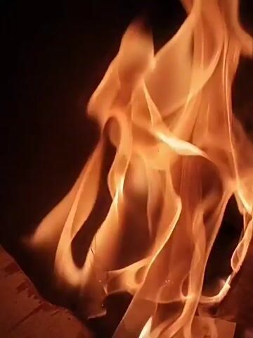 The video shows a close-up shot of flames flickering against a dark background. The flames are predominantly orange and yellow in color, with varying levels of brightness and translucence. The fire appears to be consuming wood, as hinted by visible structures at the bottom of the frame. The movement of the flames is dynamic and fluid. A faint voice can be heard in the background.