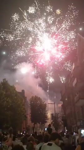 This media file captures a vibrant fireworks display in Differdange, Luxembourg. The scene is set at night, with the dark sky serving as a dramatic backdrop for the explosive pyrotechnics. A crowd of people is visible in the foreground, silhouetted against the bright lights and looking upwards in anticipation and enjoyment of the spectacle.

The fireworks are the central focus, erupting in a dazzling array of colors and patterns. We see bursts of white, pink, and red, with trails of light streaking across the sky. Smoke billows from the explosions, adding to the atmosphere. Buildings and trees line the street in the background, suggesting an urban or town setting. The overall mood appears festive and celebratory, indicative of a special occasion or holiday. The time of day is clearly night, and the weather seems clear, allowing for an unobstructed view of the fireworks. No visible text is present in the media file.