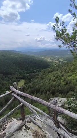 The video shows a scenic view of a lush green valley from a rocky overlook. A rustic wooden railing, made of rough-hewn logs, is in the foreground, suggesting a viewpoint or observation deck. The valley is densely covered with evergreen trees, and the terrain appears mountainous with rolling hills stretching into the distance. The sky above is partly cloudy, with patches of blue sky visible between white, fluffy clouds. The lighting suggests it is daytime, likely midday or afternoon, given the brightness and contrast. There are no people or animals visible in the video. The overall atmosphere is one of natural beauty and tranquility.