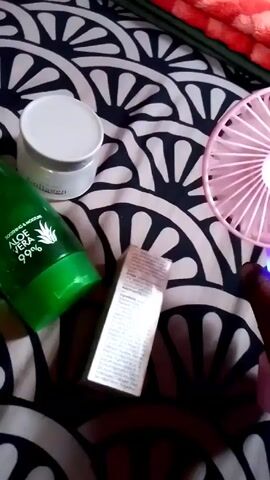 The video shows a hand holding a pink handheld fan with a purple light at the base of the handle. The fan is on and the blades are rotating. The background is a bedspread with a black and white fan-shaped pattern. There are several items on the bedspread including a green bottle labeled "Aloe Vera 99%", a white jar labeled "Collagen", and a small white box with text on it. There is a red cloth visible in the upper right corner of the frame. The camera is handheld and slightly shaky.