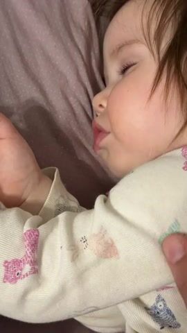 The video shows a sleeping baby with brown hair lying on their side on a mauve-colored pillow. They are wearing a cream-colored pajama top with various animal patterns in pink, blue, and brown. The baby's right hand is resting near their face. A hand briefly touches the baby's hair, then retracts. The camera remains static throughout the brief shot.