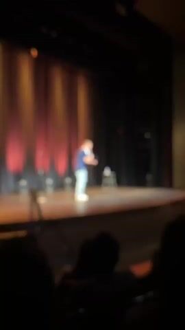 A man with light-colored hair is standing on a stage, holding a microphone. He is wearing a dark blue t-shirt with white stripes on the sleeves and light blue jeans. A stool is placed behind him on the stage. The stage has a wooden floor and is lit from the front with warm-toned lights, and there are red curtains in the background. The man is leaning forward, appearing to be speaking into the microphone. The perspective is from an audience member, with the foreground showing the blurry silhouettes of people's heads. The overall scene suggests a live performance, likely stand-up comedy.