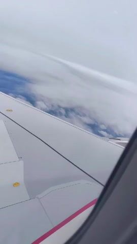 This is a first-person view from an airplane window. The camera is looking out over the wing of the plane. The wing is white with some gray sections and red and pink stripes near the bottom. There are yellow circular markings on the wing. The sky is overcast with thick white clouds. Below the clouds, glimpses of blue sky and what appears to be land or water can be seen. The camera angle is tilted upwards, suggesting the plane is in flight. There are no people or specific actions visible, only the wing and the sky.