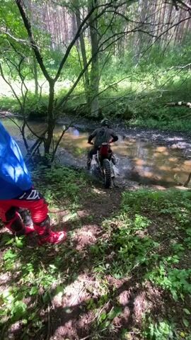 In a wooded area near Парэцкі сельскі Савет, Belarus, two individuals are engaged in off-road motorcycling. The scene is a natural setting with lush green foliage and tall trees. A stream or muddy path cuts through the terrain, and one of the motorcyclists is actively riding their dirt bike through the water. The other person, partially visible in the foreground, is standing still, dressed in similar riding gear.

The time of day appears to be daytime, with sunlight filtering through the trees, casting shadows on the ground. The overall atmosphere suggests an adventurous or recreational activity, likely enjoyed on a pleasant day, as indicated by the vibrant greenery and visible light. The presence of a stream implies a natural, possibly remote, location suitable for such pursuits. The riders are equipped with protective gear, including helmets and riding suits, highlighting the nature of the activity. No visible text is present in the media file.