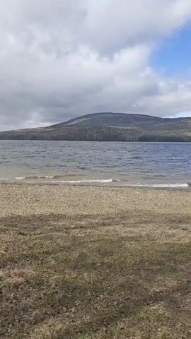 The scene is a steady, medium shot of a lake shore. The foreground is a grassy area transitioning into a pebble-covered beach. Small waves ripple across the surface of the blue lake. In the distance, a mountain range stretches across the horizon, covered with trees. The sky is partly cloudy with patches of blue. The overall mood is peaceful and serene.