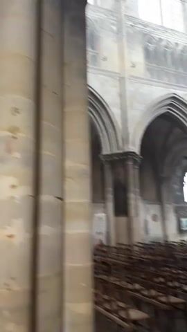 The video shows the interior of a large, stone church. Rows of wooden chairs are neatly arranged in the foreground, facing a raised altar area. The architecture is Gothic, characterized by pointed arches, ribbed vaults, and large stained-glass windows. Sunlight streams through the windows, illuminating dust motes in the air. In the background, a few people can be seen sitting in the chairs or walking, and a person is seated in a chair on the right side of the frame. Paintings and religious iconography are visible on the walls. The overall atmosphere is one of solemnity and tranquility.