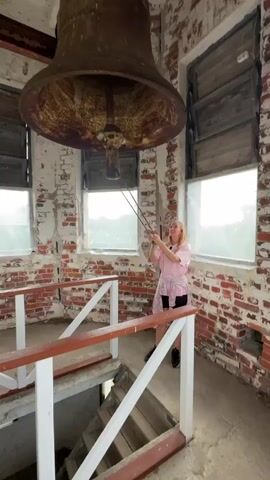 A woman is standing in a brick tower and ringing a large, rusty bell. She is pulling on a rope that is attached to the bell. The bell is hanging from the ceiling of the tower. There are windows on either side of the bell, and a set of stairs leading down from the platform where the woman is standing. The walls of the tower are made of brick and are partially covered in white plaster.