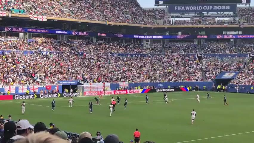 A wide shot captures a soccer match from the stands. The field is green with white lines. Several players in white and dark uniforms are visible on the field, some running and some gathered near the goal. Spectators fill the tiered stadium seats, a mass of colorful clothing. A large scoreboard in the upper right corner displays "FIFA WORLD CUP 2026". Banners and advertisements are visible around the stadium, including "Globaant" and "Betano". The atmosphere appears lively, with many people in attendance.
