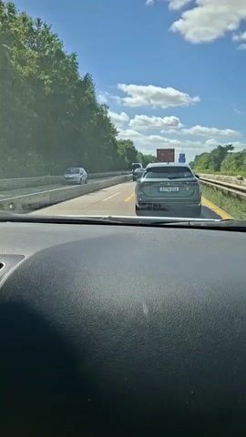 A first-person perspective video shows a traffic jam on a highway. The camera is positioned inside a car, facing forward through the windshield. The road is a multi-lane highway with a clear blue sky and scattered clouds overhead. Trees line the sides of the highway. In the foreground, the dashboard of the car is visible. In the middle ground, traffic is backed up, with several cars and trucks stopped. A dark green SUV is directly in front of the camera car. To the left, another car is visible. Further ahead, traffic continues to be congested. The overall impression is of a slow-moving or stopped commute.