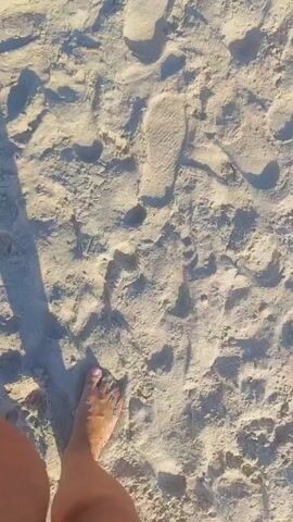 The video shows a first-person perspective of someone walking on a sandy beach towards the ocean. Footprints are visible in the sand. The person's legs and feet, with painted toenails, are in the lower left of the frame. Shadows of the person and possibly another individual are cast on the sand. The ocean with gentle waves and a clear blue sky are visible in the background, with a few distant figures in the water.