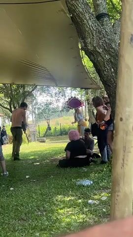 The video shows a group of people outdoors, possibly at a festival or gathering. The foreground is dominated by green grass, with some scattered plastic bottles. A large, light-colored tarp is stretched overhead, providing shade. Behind the tarp, several individuals are visible. In the center, a shirtless person is holding a red and gold drum aloft, seemingly in the process of hitting it. To their left, another person in a blue top appears to be dancing or moving rhythmically. To the right, a person in a black bikini top and denim shorts is holding a drink and looking towards the center. Another individual, with pink hair and wearing a black top, is seated on the grass in the middle ground. The background consists of trees and foliage, suggesting a natural, possibly wooded, setting. The lighting indicates it is daytime, with bright sunlight filtering through the leaves. The overall atmosphere appears relaxed and energetic.
