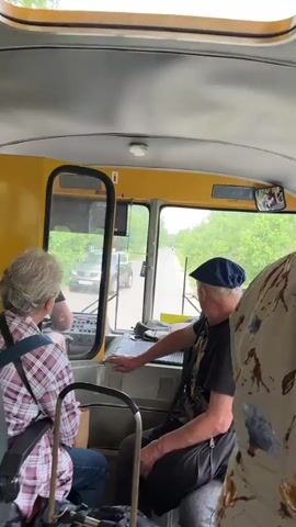 The video shows the interior of a yellow bus, looking forward towards the driver's cabin and the road ahead. Several passengers are visible. In the foreground, the back of a person wearing a floral dress is seen on the right. To the left, a person with gray hair wearing a plaid shirt and blue jeans is seated, facing forward. Next to them, a person wearing a dark shirt, dark pants, and a blue cap is seated and appears to be interacting with the driver or the bus controls. The driver is partially visible through the window of the cabin. Luggage, including a wheeled shopping bag and a green bag, is present on the floor. The bus is on a paved road surrounded by greenery, suggesting a rural or semi-rural setting. The lighting indicates it is daytime.