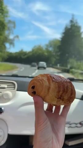The video shows a person holding a pain au chocolat in a car. The car appears to be a Fiat 500 based on the dashboard and logo. The person is holding the pastry in their right hand, in front of the dashboard. In the background, the view through the windshield shows a road with trees and other cars. The sky is blue with some clouds. The video seems to be recorded from the passenger seat.