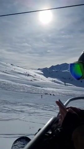 The video depicts a view from a ski lift ascending a snowy slope on a sunny day. In the foreground, an empty ski lift chair swings gently. Below, the snow-covered terrain reveals tracks made by skiers and snowboarders. Several individuals can be seen skiing down the slope, dispersed across the field. The sky above is a mix of blue and patches of cloud, with the sun shining brightly. In the distance, snow-capped mountains are visible against the horizon. The overall atmosphere is bright and the scene captures the essence of a day at a ski resort.