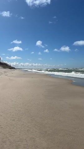The video shows a wide, sandy beach with gentle waves rolling in. The sky is blue with scattered, fluffy white clouds. There are no people visible on the beach. The water is a dark green color. There is some vegetation on the left side of the frame in the distance.