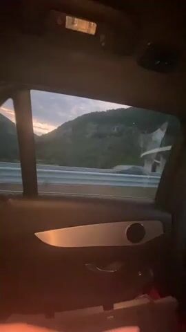 The video shows the interior of a car with a window view of a highway and hills. In the foreground, a dog is visible in a carrier. The dog is dark brown with large ears and appears to be lying down or resting. The car is in motion, as indicated by the blurred scenery outside the window and the movement within the car. The lighting suggests it is either dusk or dawn.