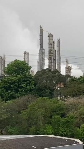 The video shows an industrial complex with numerous tall towers, some of which are emitting plumes of white smoke against a cloudy sky. The complex is surrounded by lush green trees, with some residential buildings visible among the foliage. The camera pans slowly from left to right, revealing more of the industrial structures and the surrounding vegetation. The overall impression is one of an industrial area nestled within a natural environment.