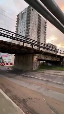 A handheld video pans under an overpass. The overpass is made of concrete and metal beams. The concrete supports of the overpass are covered in graffiti. Beyond the overpass, a tall building under construction is visible. Construction equipment, including an excavator, is present on the left side of the frame. The sky appears overcast.