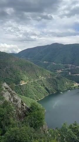 A vertical video captures a panoramic view of a vast, dark blue lake nestled within a valley of lush, green mountains under a cloudy sky. The camera pans slightly to the right, revealing winding roads carved into the mountainsides. Several small white boats are visible on the water, some clustered together and others further apart. In the distance, a few small buildings appear on a peninsula jutting into the lake, and another is situated on a small island. The foreground features a rocky cliff face with sparse green foliage. The overall scene is serene and showcases the natural beauty of the landscape.