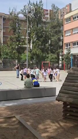A group of children and one adult are playing in a schoolyard. The children are holding hands in a circle and appear to be dancing or playing a game. Two children are sitting on a concrete ledge watching the others. The schoolyard has a sandpit, a wooden play structure, and a soccer goal in the background. The buildings in the background appear to be apartment buildings. The weather is sunny.