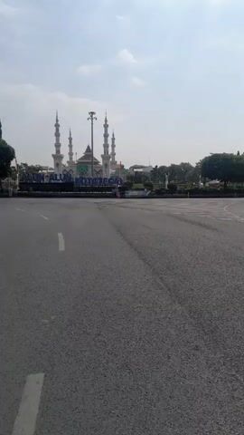 A horizontal video shows a mosque with multiple minarets in the background, along with a sign that reads ALUN-ALUN KOTA TEGAL. The foreground is a street with white lane markings. Trees and a flagpole are visible to the left and right of the mosque. The sky is cloudy.