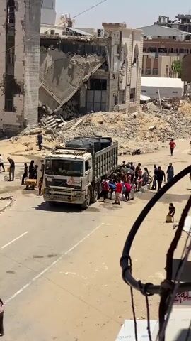 This media file captures a scene of apparent humanitarian aid distribution in Khan Yunis, Palestinian Territories. The primary subjects are a group of people, including men, women, and children, gathered around a large white truck. The truck itself is a significant object, with its bed filled with what appear to be large black water tanks. It bears a logo on its side that reads "Médecins Sans Frontières" (Doctors Without Borders), indicating its role in providing essential supplies.

The activity taking place is the distribution of resources, likely water, from the truck to the people. Many individuals are seen holding buckets and containers, some of which are filled. The people appear to be actively engaged in collecting and carrying these containers. There is also a person in a wheelchair present, suggesting the aid is being distributed to a diverse group of recipients.

The scene is set on a dusty, sun-baked street, with the backdrop of partially destroyed buildings. The rubble and damage to the structures suggest a recent event of destruction, possibly conflict. The bright sunlight and clear skies indicate it is daytime, likely mid-morning or early afternoon. The emotions conveyed are a mixture of necessity and perhaps a sense of relief, as people work together to obtain vital resources. The overall atmosphere is one of a community in need, relying on external aid.

Visible text on the truck includes the "Médecins Sans Frontières" logo, which is in both French and Arabic script, and a license plate that appears to have Arabic characters. The overall impression is one of resilience and the ongoing efforts to support a population affected by hardship and destruction.