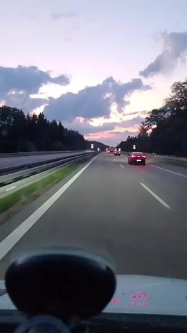 The video is a first-person view from inside a car driving on a highway at dusk. The sky is a gradient of purple and pink with clouds. Trees line both sides of the highway, which has multiple lanes. Several cars are visible on the road, including a red sports car in the right lane. The dashboard of the car is partially visible in the foreground, with a dark, round object in the lower left corner and red lights on the dashboard. Traffic signs are visible on the side of the road. The lighting suggests it is either sunrise or sunset, with the sun below the horizon.