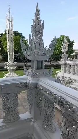 The video pans across an ornate white structure with intricate detailing. In the foreground, a white railing with elaborate carvings is visible. Beyond the railing, a lush green lawn stretches out, bordered by trees. Two tall, spire-like white structures stand in the distance. The architecture features sharp, upward-reaching elements and detailed decorative patterns. The sky is partly cloudy. The camera movement is smooth, offering a continuous view of the scene.