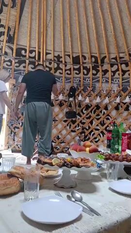 A group of people are gathered inside a yurt, a traditional nomadic dwelling. The yurt is circular with wooden lattice walls and a conical roof, decorated with patterned fabrics and hanging tassels. A table is laden with a feast, including grilled fish, bread, fruits, and bottles of beverages. Two individuals are visible near the entrance, one standing and another bending down to attend to something on the floor. The scene suggests a meal or gathering is about to commence or is in progress.